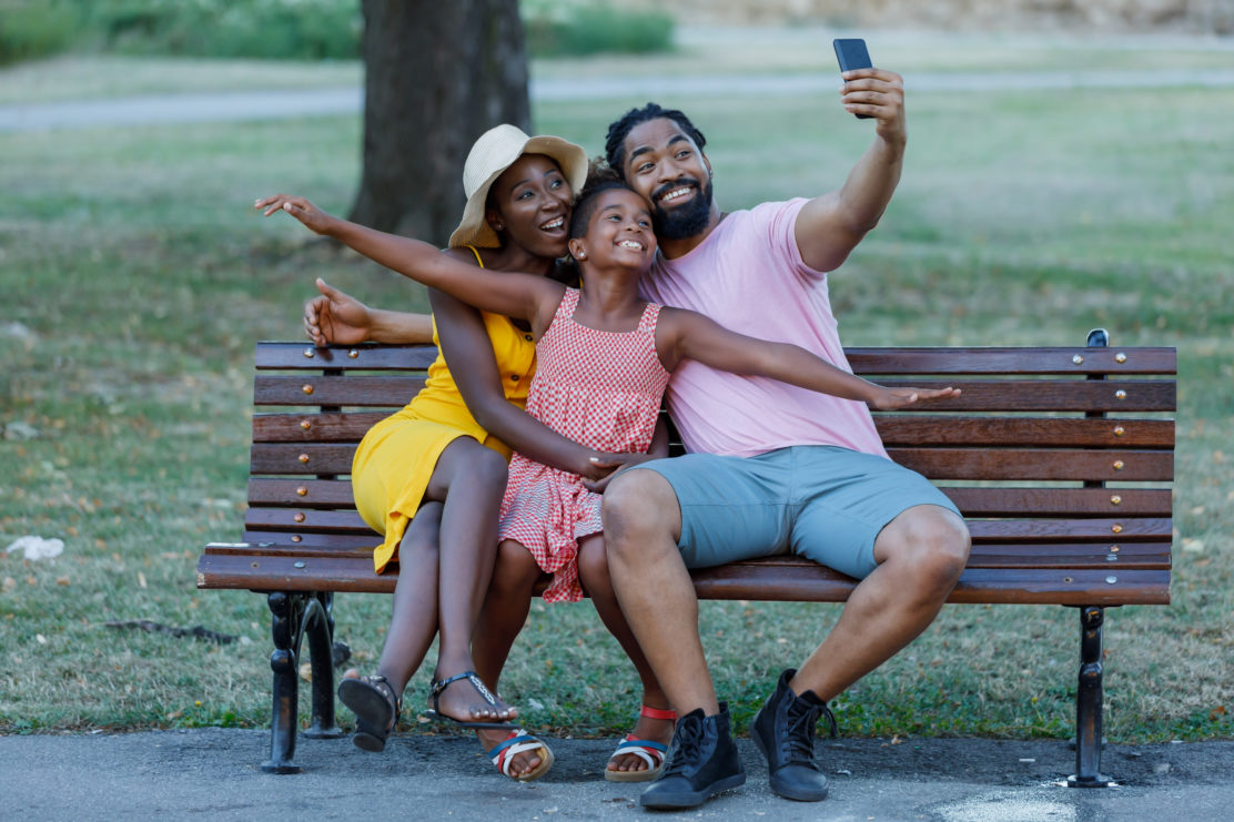 Young and happy family is enjoying the time together, sitting on a bench in a public park and taking a selfie